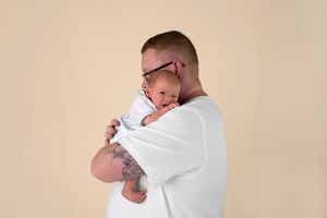 Tattooed father with glasses holding sleeping newborn baby wrapped in white blanket against beige background.