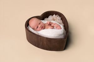 Newborn baby swaddled in white wrap sleeping in a heart-shaped wooden bowl on a beige background.