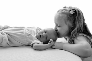Black and white photo of a young girl gently kissing a newborn baby lying on a soft white surface.