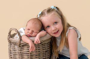 A young girl with blue hair bows smiles beside a sleeping newborn baby resting in a wicker basket.