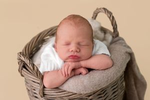 Peaceful newborn baby sleeping in a rustic wicker basket with soft gray blanket, wearing white onesie.