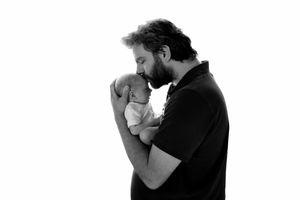 Black and white photo of a bearded father tenderly holding and kissing his newborn baby against white background.