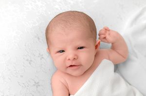 Adorable newborn baby lying on white blanket, looking up with bright eyes and tiny fist raised.