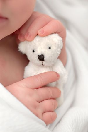 Newborn baby hands gently holding a tiny white teddy bear while wrapped in a soft white blanket.