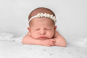 Sleeping newborn baby girl wearing a white floral headband, resting chin on hands on a white lace surface.