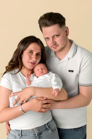 Young couple in white shirts holding their newborn baby against a beige studio background.