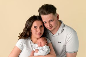A couple poses with their newborn baby against a beige background in a professional studio portrait photo.