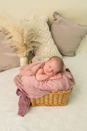 Sleeping newborn baby wrapped in pink muslin cloth nestled in a wicker basket with boho pillows and pampas grass decor.