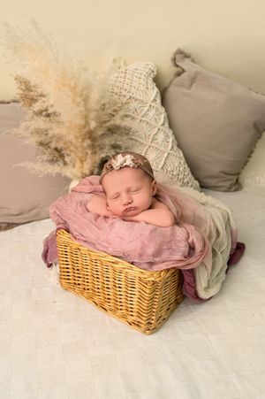 Sleeping newborn baby in a wicker basket with pink wrap and floral headband, surrounded by soft pillows and pampas grass.