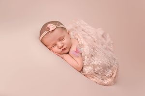 Newborn baby girl sleeping peacefully wrapped in pink lace swaddle with floral headband on beige background.