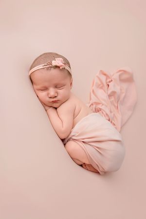 Newborn baby girl sleeping peacefully wrapped in pink fabric with a floral headband on a pink background.