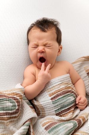 Newborn baby yawning with hand near mouth, wrapped in a colorful patterned blanket on a white background.