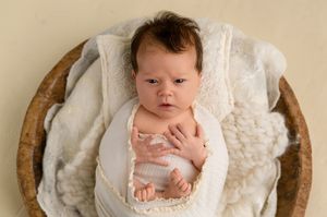 Newborn baby swaddled in white wrap, resting in a wooden bowl with soft cream blankets.