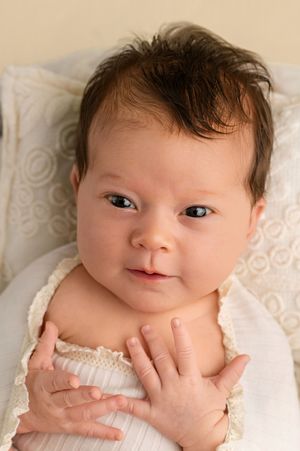 Newborn baby with dark hair and blue eyes lying on white blanket, hands folded on chest, looking at camera.