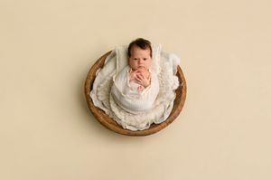 Newborn baby wrapped in white swaddle cloth resting in a round wooden bowl on a cream background.