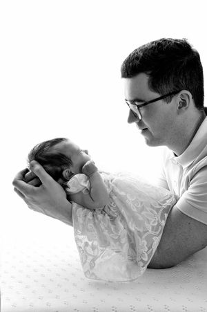 Black and white photo of a father in glasses tenderly holding his sleeping newborn baby in both hands.