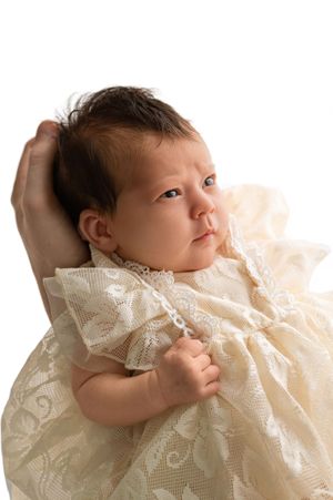 Newborn baby in cream lace christening gown being cradled by adult hands against white background.