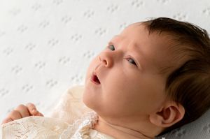 Newborn baby with dark hair lying on white textured blanket, looking upward with curious blue eyes.