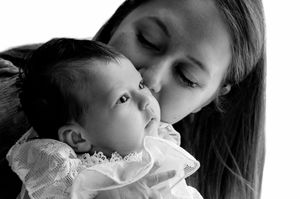 Black and white photo of a young woman tenderly kissing a newborn baby dressed in white lace.