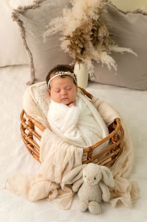Newborn baby girl swaddled in white wrap sleeping in wicker basket with bunny toy and pampas grass decor.