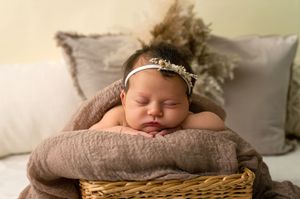 Sleeping newborn baby girl in floral headband resting in wicker basket with soft brown wrap.