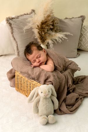 Sleeping newborn baby in a wicker basket with a gray blanket, bunny stuffed animal, and decorative pillows.