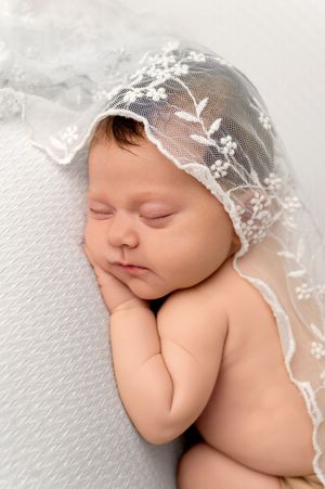 Sleeping newborn baby wearing a delicate white lace bonnet with floral embroidery, resting peacefully on a soft white surface.