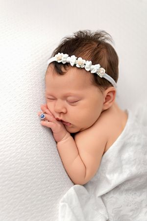 Sleeping newborn baby girl wearing a white floral headband, wrapped in a soft white blanket on a white background.