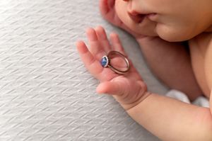 Newborn baby hand holding a delicate sapphire engagement ring on a white textured surface.