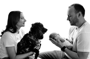 Black and white family portrait with mother holding dog, father cradling newborn baby, wearing white shirts.