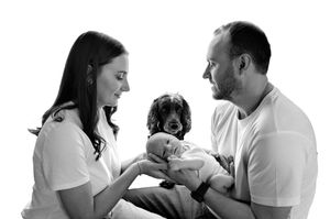 Black and white family portrait of parents holding newborn baby with a cocker spaniel dog between them.