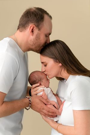 New parents sharing a kiss over their newborn baby, both dressed in white, in a tender family portrait.