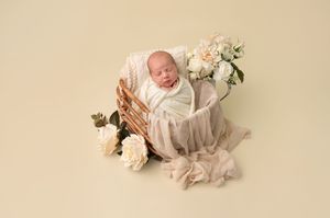 Newborn baby wrapped in cream swaddle sleeping in wicker basket with white roses on beige background.