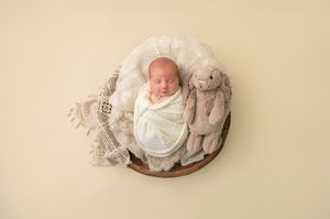 Newborn baby swaddled in white wrap sleeping in wooden bowl with stuffed bunny on cream background.