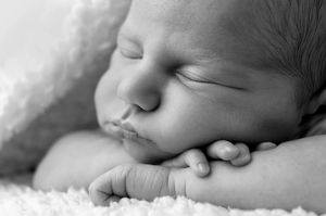 Close-up black and white photo of a sleeping newborn baby with hands tucked under cheek on soft blanket.