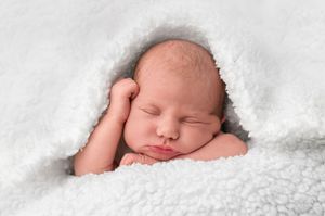 Peaceful newborn baby sleeping wrapped in soft white fluffy blanket with tiny hand tucked under cheek.