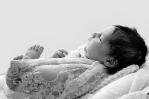 Black and white photo of a sleeping newborn baby in lace outfit, lying peacefully on soft white blanket.