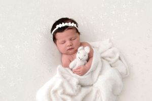 Newborn baby sleeping wrapped in white blanket, holding a small bunny toy, wearing a floral headband.