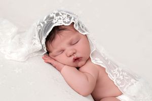 Sleeping newborn baby wearing a delicate white lace bonnet, posed peacefully on a soft white backdrop.