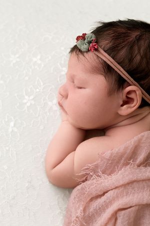 Sleeping newborn baby girl wearing a pink floral headband, wrapped in a soft blush wrap on a white lace backdrop.