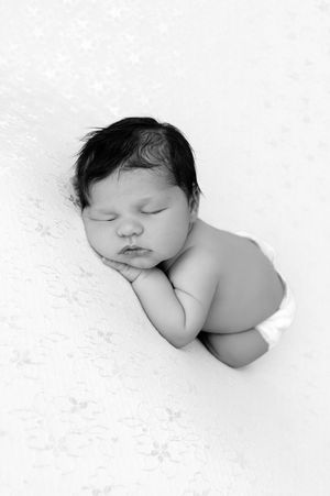 Black and white photo of a sleeping newborn baby with dark hair, curled up peacefully on a soft white blanket.