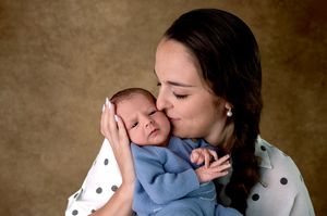 A loving mother gently kisses her newborn baby wrapped in a soft blue blanket during a studio portrait session.