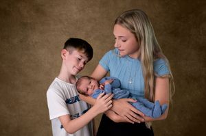 Two siblings, a boy and a teenage girl, lovingly hold a newborn baby dressed in blue during a studio portrait session.