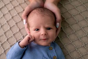 Newborn baby in blue knit outfit lying on blanket, cradled gently by adult hands in a serene portrait.