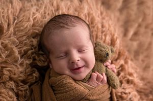Smiling newborn baby wrapped in golden knit swaddle holding a tiny teddy bear on a fluffy brown backdrop.