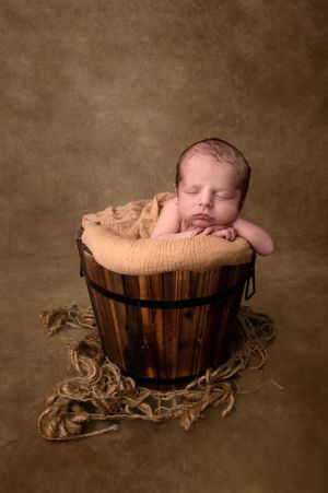 Sleeping newborn baby nestled in a rustic wooden bucket with burlap wrap on a warm brown studio background.