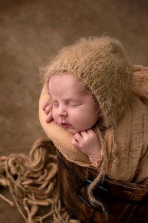Sleeping newborn baby in a wooden bucket, wrapped in a soft tan knit blanket and bonnet, posed for a professional photo.