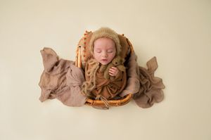 Sleeping newborn baby posed in wicker basket with soft brown wraps and bonnet.
