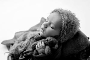 Black and white portrait of a sleeping newborn baby in knit bonnet cuddling a tiny teddy bear.