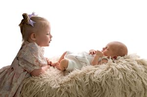 Toddler girl in floral dress gazes at newborn baby lying on fluffy white blanket in studio photo.